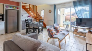 Una sala moderna con un sofá, dos sillones, una mesa de comedor con sillas, una zona de cocina con un refrigerador, y una escalera que lleva a otro nivel.