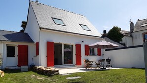Une maison avec un toit en pente, des volets rouges, une terrasse avec une table et des chaises, et un chemin de pierre menant à l’entrée.