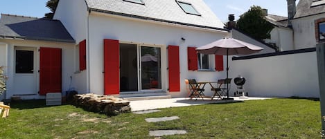 A house with a sloped roof, red shutters, a patio with a table and chairs, and a stone pathway leading to the entrance.