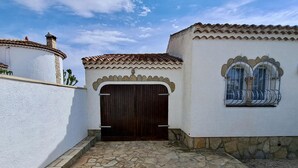 A courtyard with a stone floor, a large wooden gate, and a white building with a tiled roof and a window with metal bars. - Spacious Villa in Miami Platja (Miami Playa)