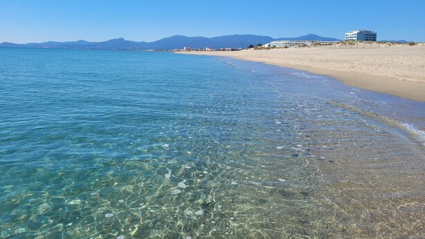 A beach with clear water, a sandy shore, and a building in the background. - Renovated Studio - 5 Pers - Sea View - Canet-en-Roussillon (Canet-en-Roussillon)