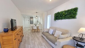 Ein modernes Wohnzimmer mit einer Couch, einem Fernseher auf einer Holzkommode, einem Essbereich mit Tisch und Stühlen sowie einer Küche im Hintergrund.