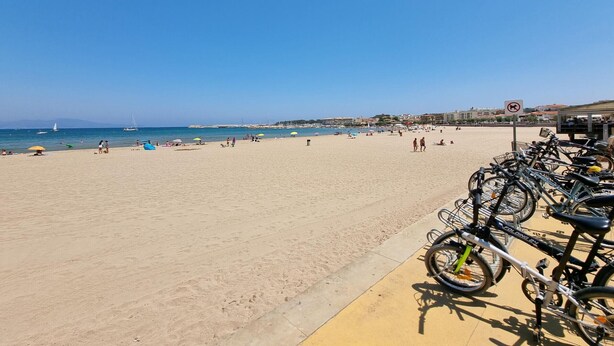 Ein Strand mit einer Reihe Fahrrädern an der rechten Seite, einem klaren blauen Himmel und einem Sandstrand mit Menschen und Sonnenschirmen.
