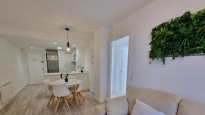 Ein modernes Wohnzimmer mit einem beigefarbenen Sofa, einem Esstisch mit Stühlen, einer Küchenzeile und einer grünen Wanddekoration.