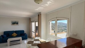 A living room with a blue sofa, a coffee table, and a dining area with a white table and chairs. There is a wooden cabinet and a window with a view of a mountainous landscape. - Charming 2-bedroom apartment with terrace and parking in Roses (Roses)