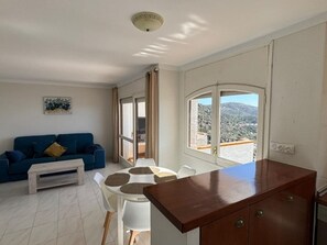 A living room with a blue sofa, a coffee table, and a dining area with a white table and chairs. There is a wooden cabinet and a window with a view of a mountainous landscape. - Charming 2-bedroom apartment with terrace and parking in Roses (Roses)