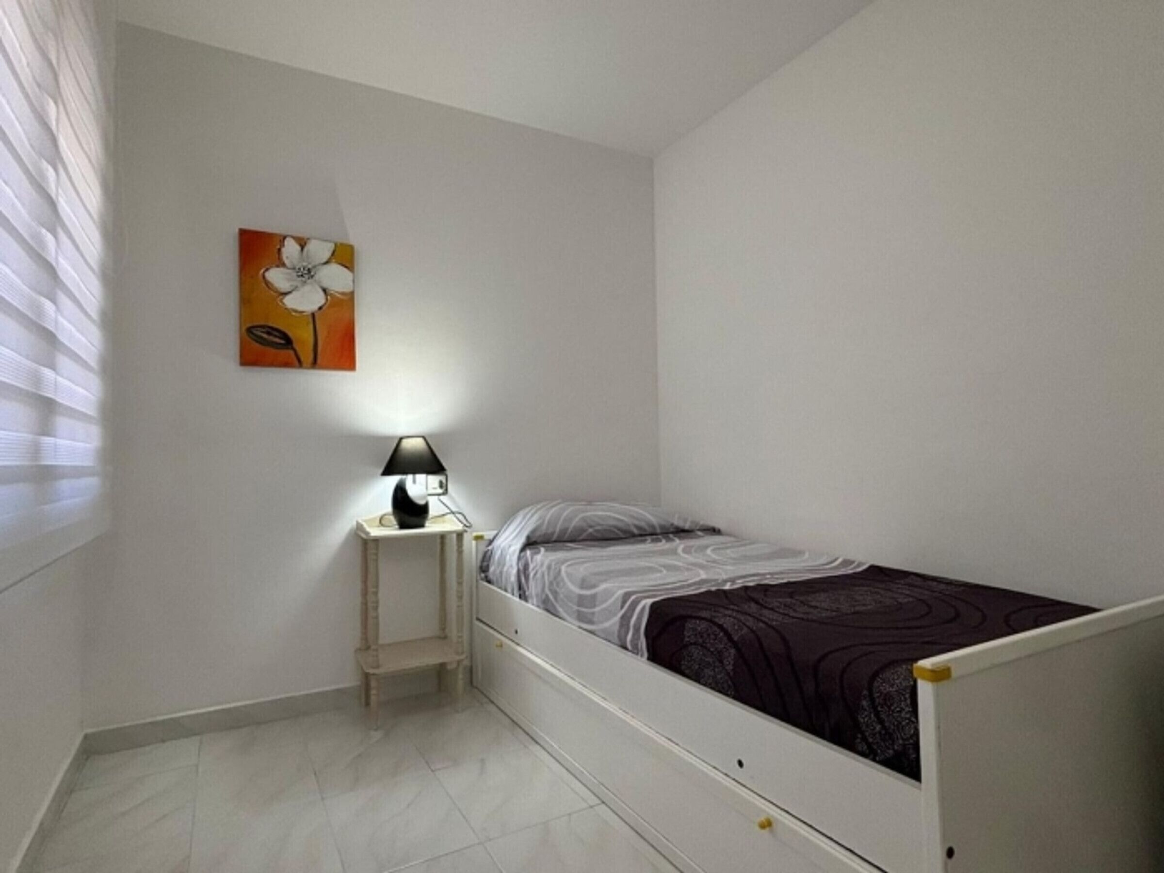 Une petite chambre avec un lit simple, une table de chevet, une lampe et un tableau au mur.