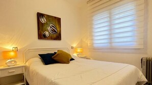 A bedroom with a large bed, two bedside tables, a wall-mounted artwork, and a window with blinds. - Charming 2-bedroom apartment with terrace and parking in Roses (Roses)