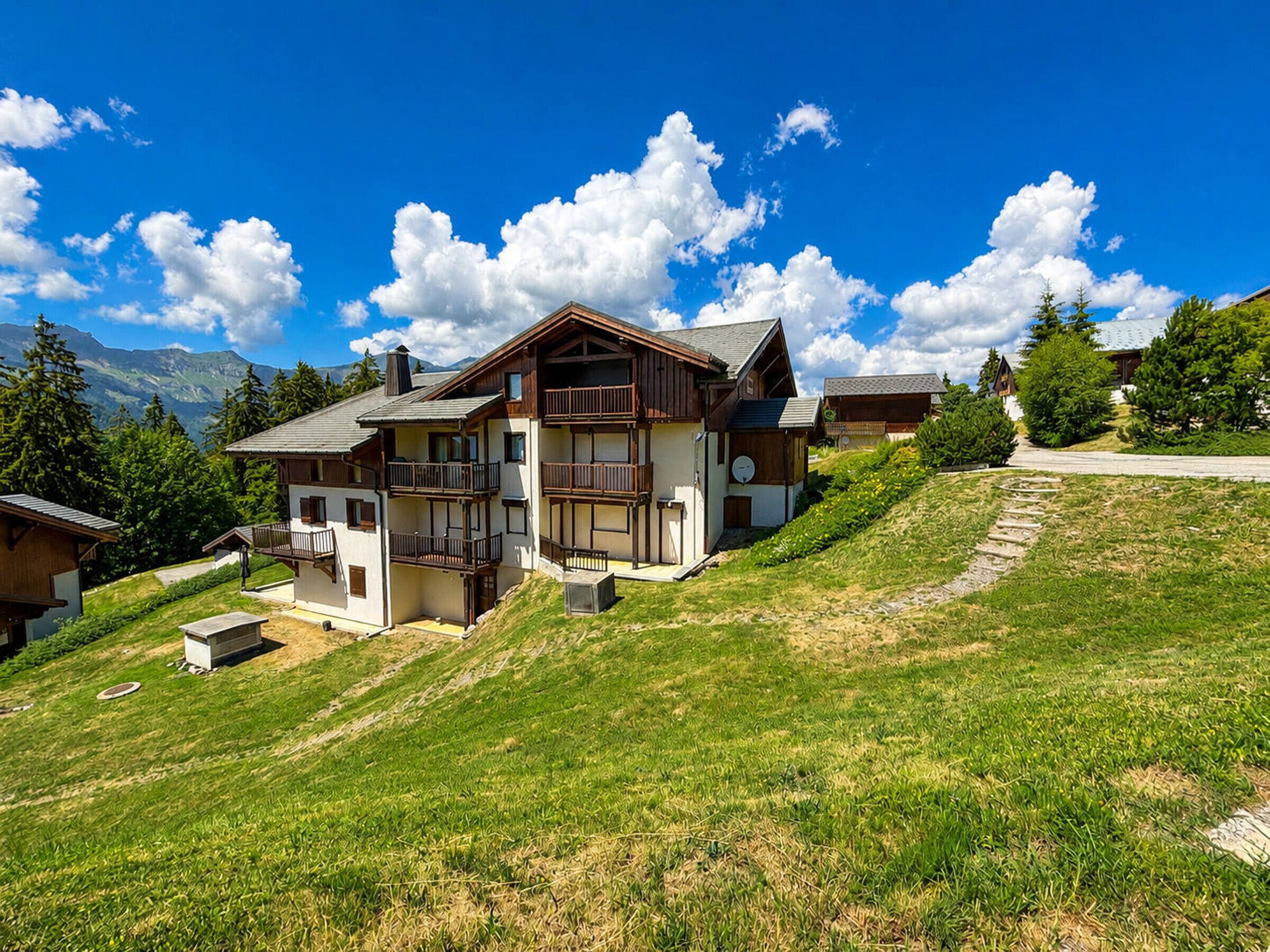 A two-story chalet with balconies, a stone pathway, and a mountainous backdrop.