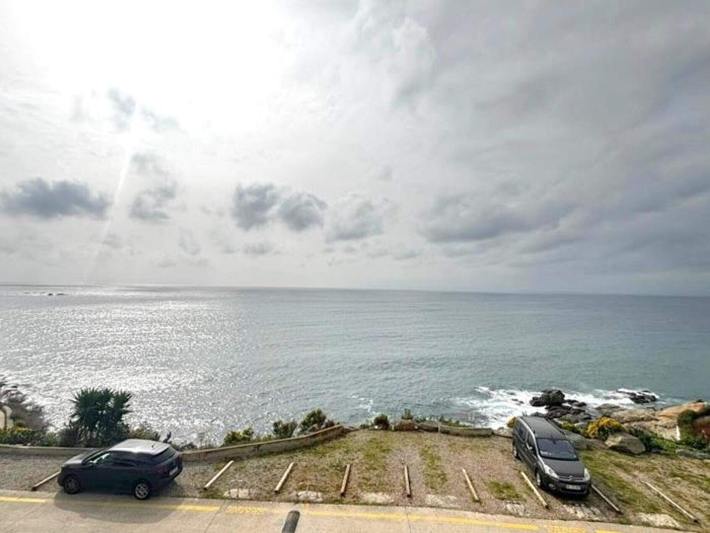 En kystnær parkeringsplads med biler, en græsklædt skrænt og udsigt til havet under skyet himmel.
