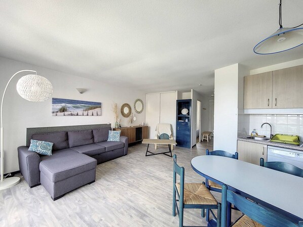 A modern living room with a grey sofa, a dining table with chairs, and a kitchen area with cabinets and a sink.