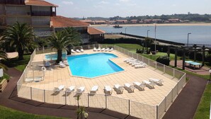 Un área de piscina con sillones de descanso, vista despejada a la playa y un edificio de techo rojo.