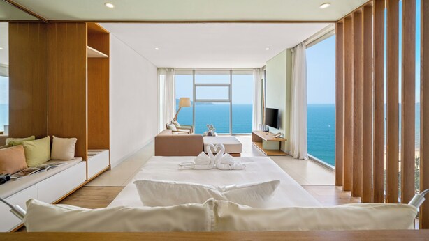 A modern hotel room with a large bed, a seating area, and a view of the ocean. - Signature Luxury Village (Da Nang)