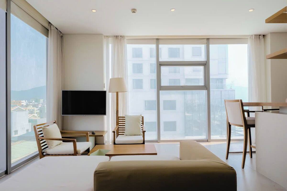 A modern hotel room with a large window, a flat-screen TV, a sofa, a dining table, and two chairs.