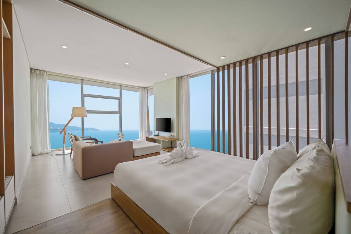 A modern hotel room with a large bed, a sofa, a desk with a TV, and a balcony with a view of the sea.