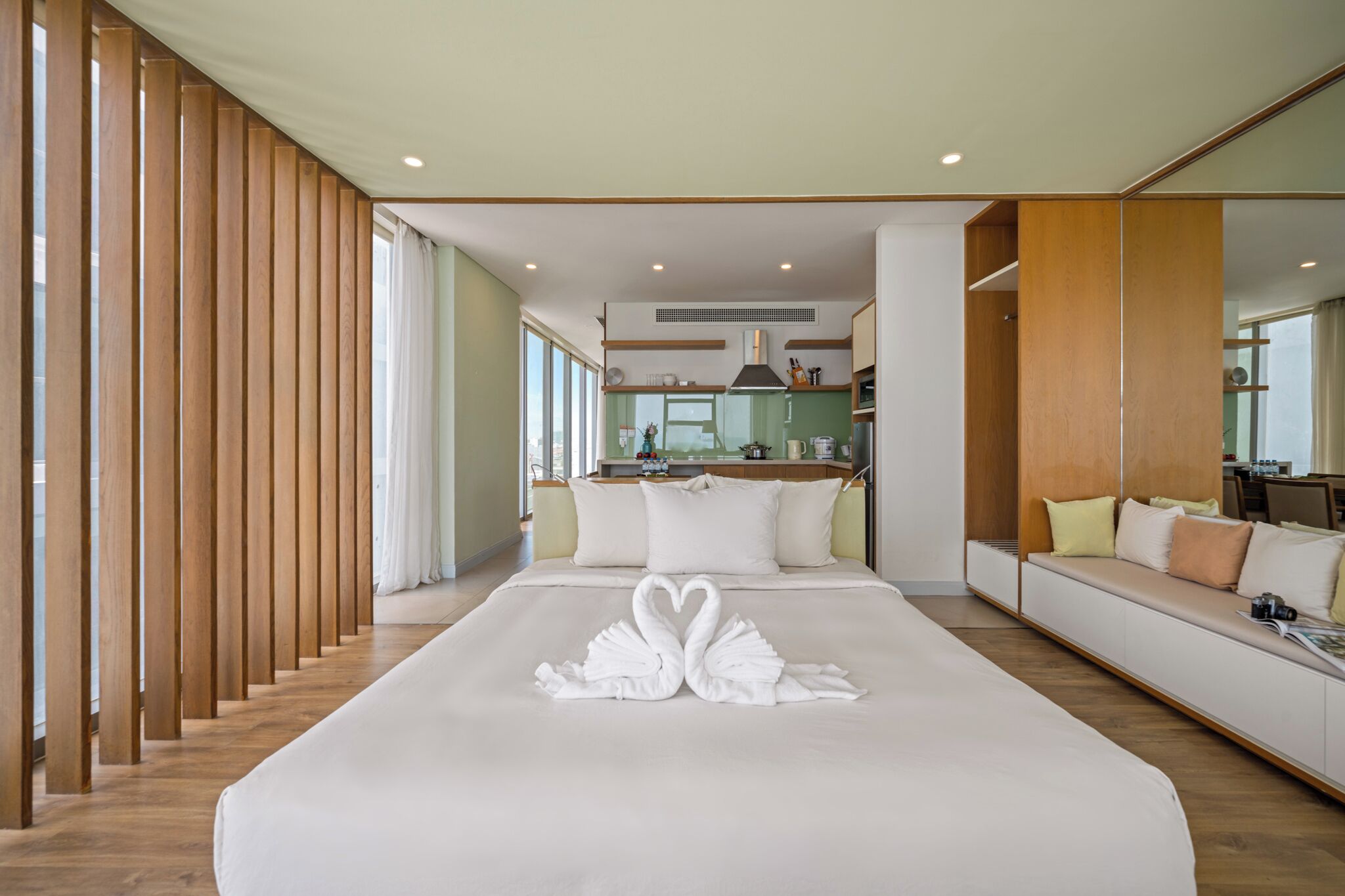A modern hotel room with a large bed, a wooden partition, a sofa, and a view of the kitchen.