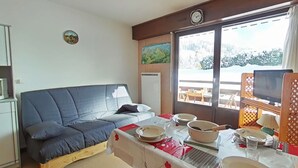 A cozy room with a bed, a dining table set for two, a television, and a window with a view of a snowy landscape. - Studio near slopes, balcony with Aravis view (Le Grand-Bornand)
