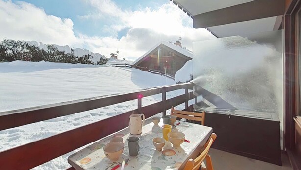 A balcony with a table set for tea, overlooking a snowy landscape and a building with a sloped roof. - Studio near slopes, balcony with Aravis view (Le Grand-Bornand)