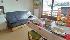 A cozy room with a sofa, a dining table set with plates, a TV, and a window with a view of the outdoors. - Studio near slopes, balcony with Aravis view (Le Grand-Bornand)