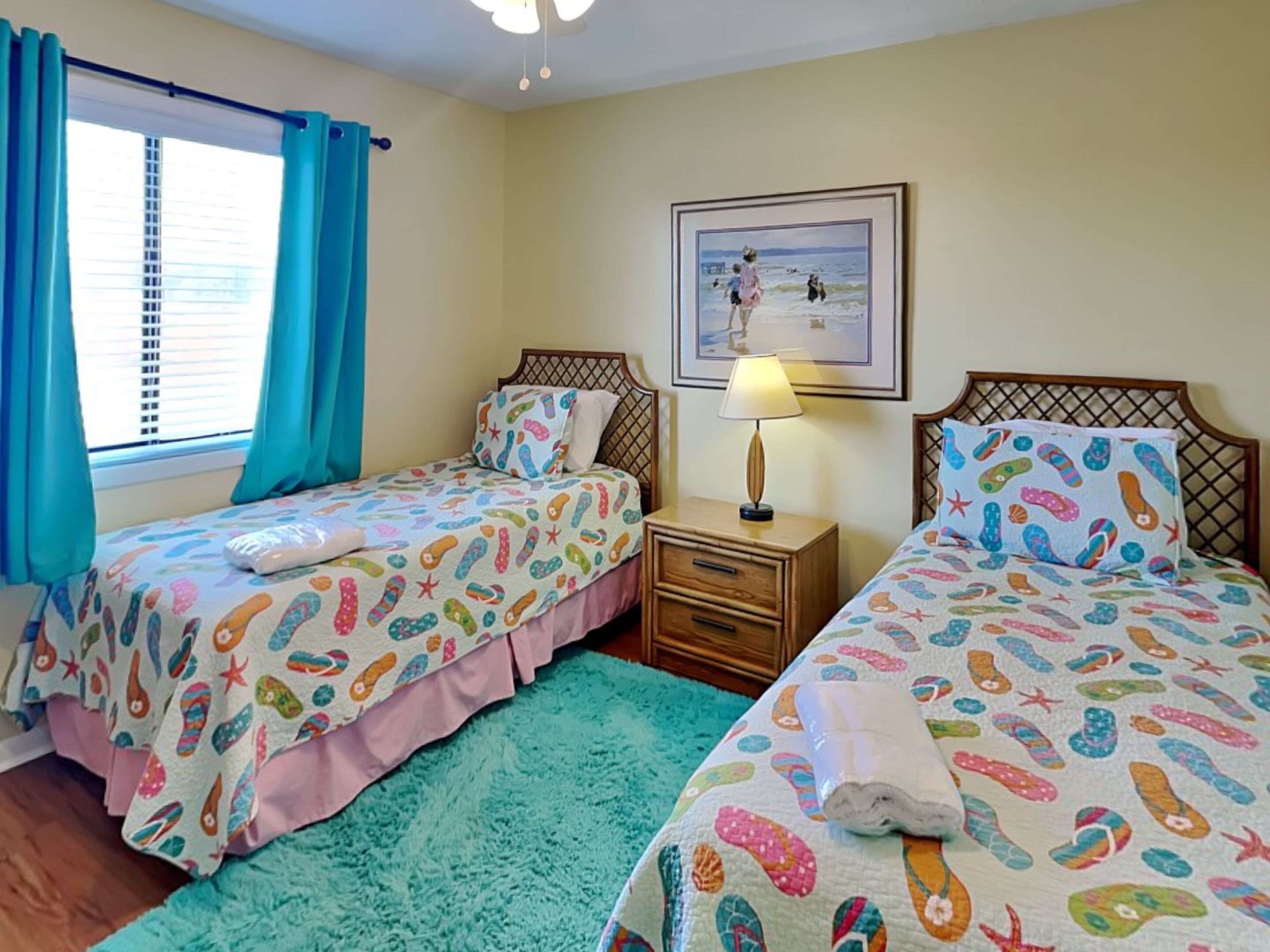 A bedroom with two beds, each with a colorful sea-themed bedspread. There is a window with blue curtains, a nightstand with a lamp, and a framed picture on the wall.