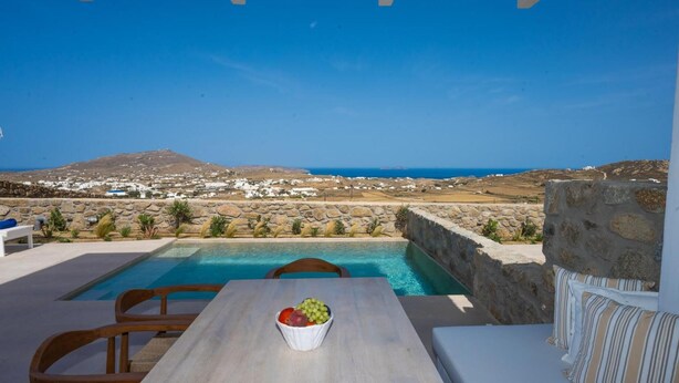 A poolside area with a table and chairs, a bowl of fruit, and a view of the sea and distant landscape. - Sunrise Villa Sea View up to 4 pax (Ano Mera)