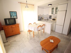 Una cocina compacta con armarios blancos, una mesa de comedor con sillas, una pequeña isla y un televisor sobre un mueble de madera.