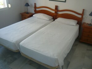 Una cama doble con cabecera y piecera de madera, dos mesitas de noche con lámparas, y suelo de baldosas.
