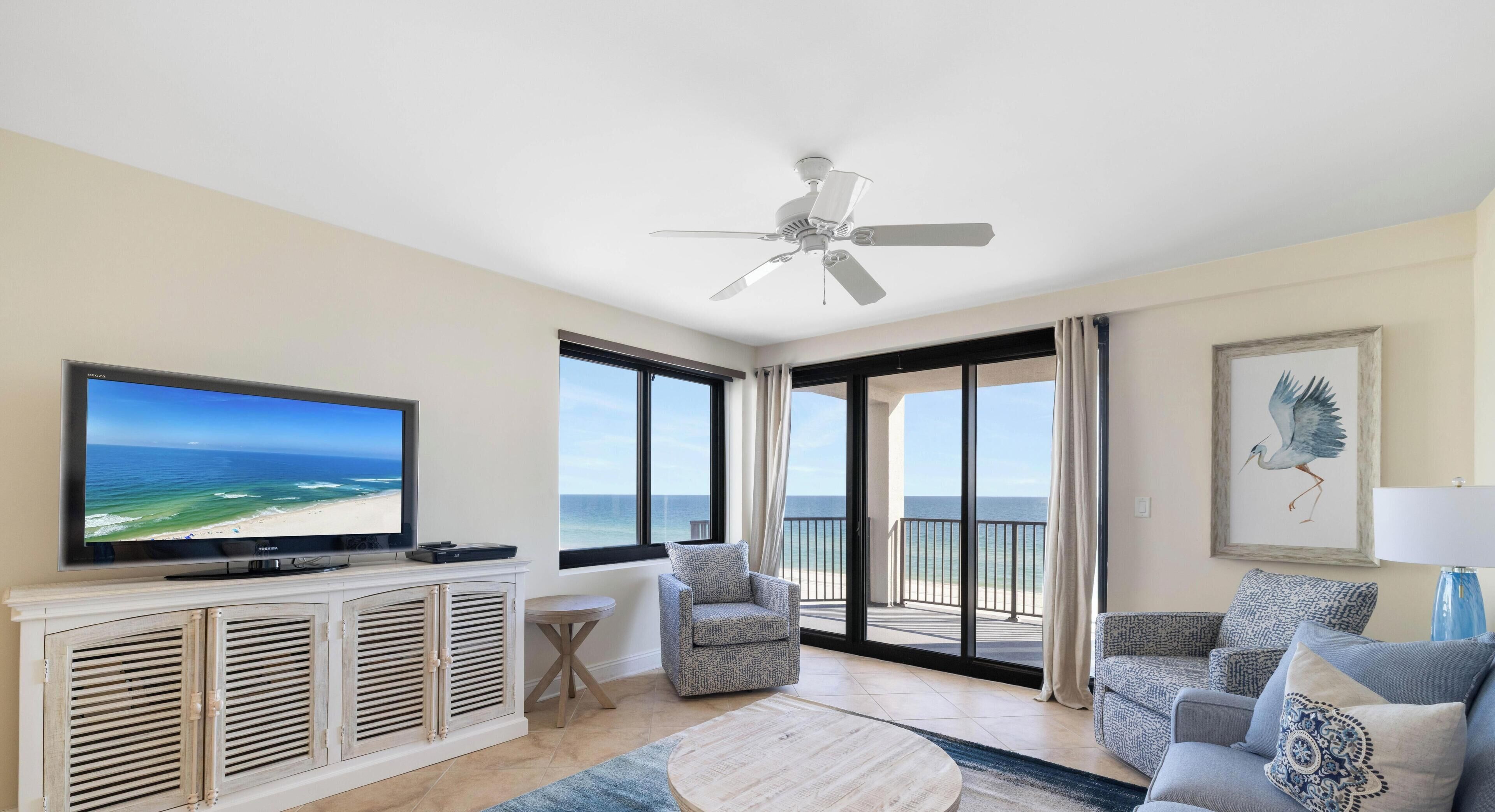 A spacious living room with a large flat-screen TV, a ceiling fan, and a balcony with a view of the ocean.