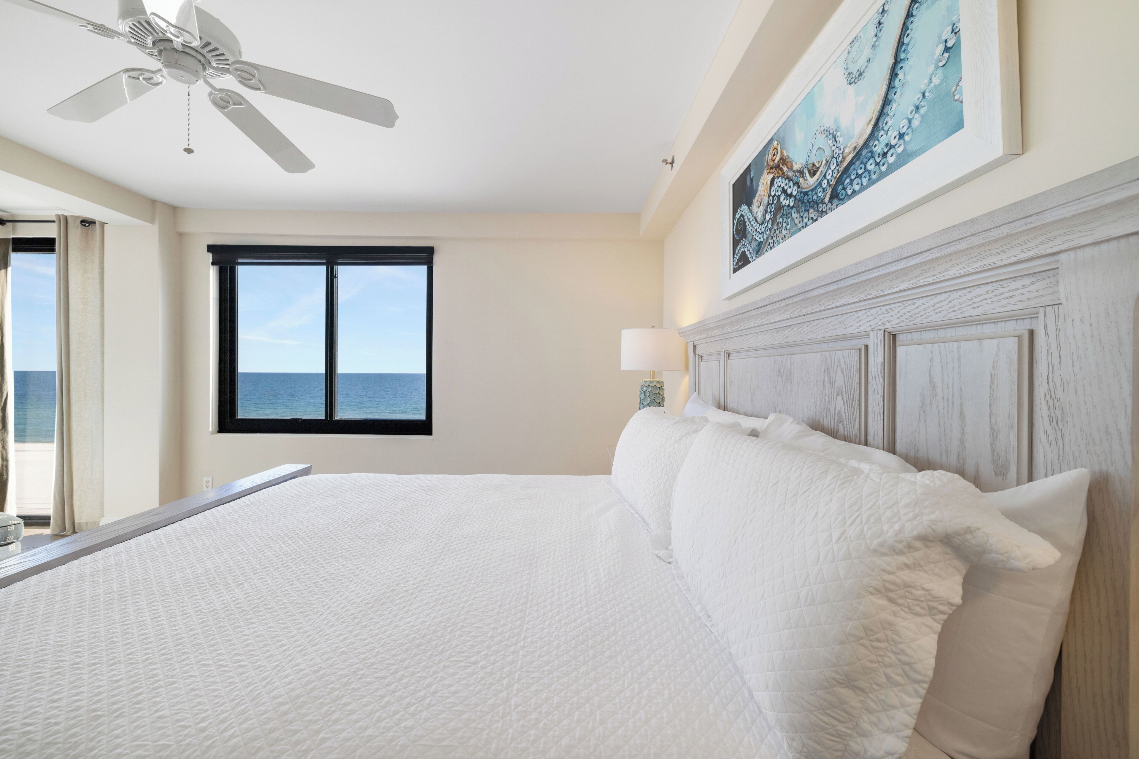 A large bed with white bedding, a ceiling fan, a window with a view of the ocean, and a painting on the wall.