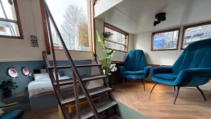 Ein Zimmer mit einer Holztreppe, einem Bett, zwei blauen Stühlen und Blick auf die Straße durch die Fenster.