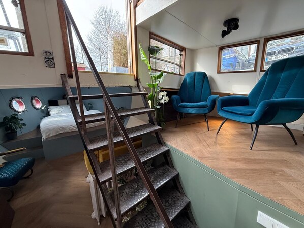 Une pièce avec un escalier en bois, un lit, deux chaises bleues et une vue sur la rue par les fenêtres.