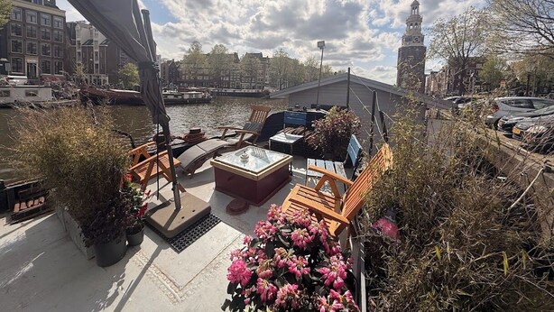 Ein Bootsdeck mit Sitzgelegenheiten und einem Tisch, Blick auf einen Fluss sowie ein Glockenturm im Hintergrund.
