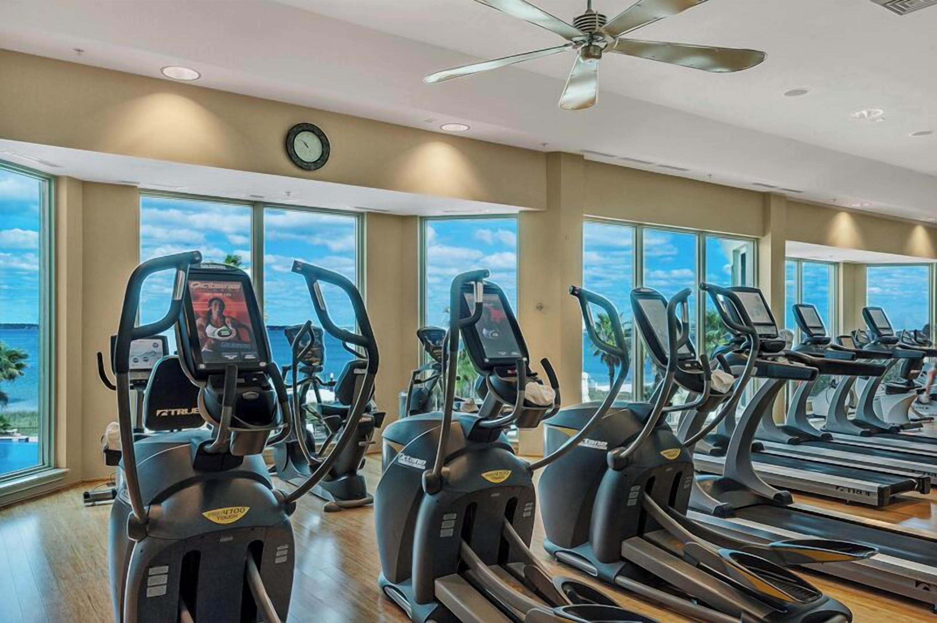 Une salle de sport équipée de plusieurs tapis de course, de grandes fenêtres et d’un ventilateur de plafond.