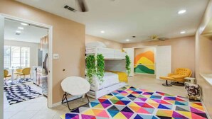 Ein Zimmer mit einem Etagenbett, einem runden weißen Stuhl, einem farbenfrohen geometrischen Teppich, einem gelben Ohrensessel und einer Tür zu einem anderen Raum.