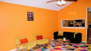 Ein Wohnzimmer mit orangefarbenen Wänden, einem schwarzen Sofa, zwei gelben Ohrensesseln, einem farbenfrohen geometrischen Teppich und einem Deckenventilator.