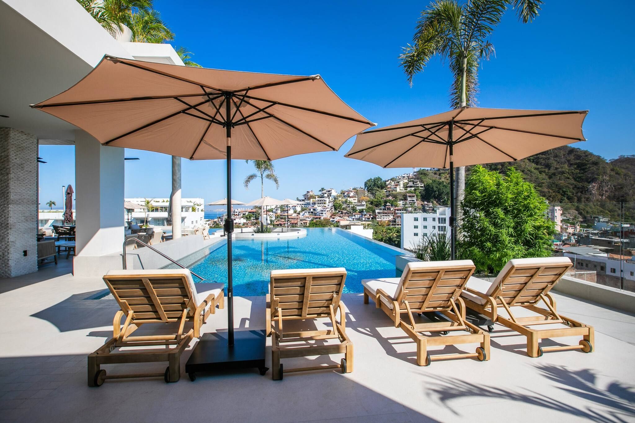 Zona junto a la piscina con sillones de descanso y sombrillas, con vistas a una zona residencial y un cuerpo de agua.