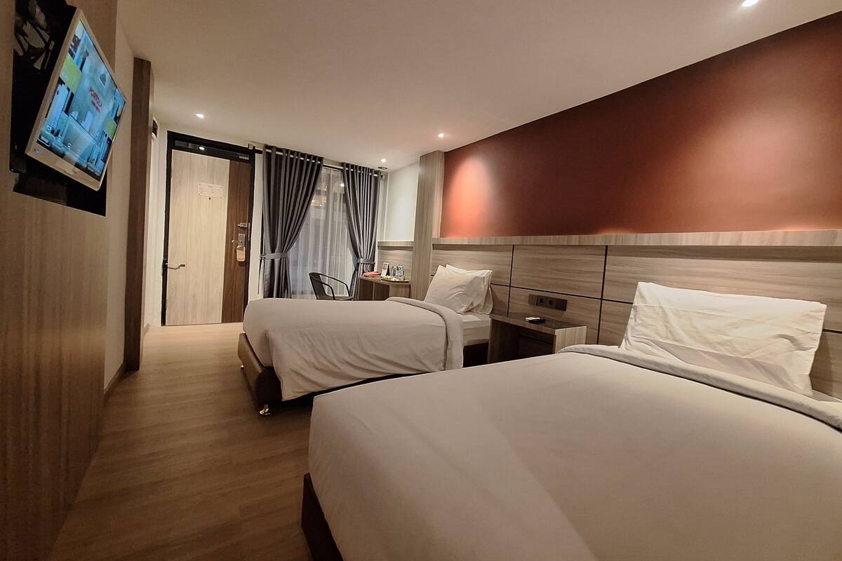 A hotel room with two beds, a TV mounted on the wall, a desk with a chair, and a window with curtains.