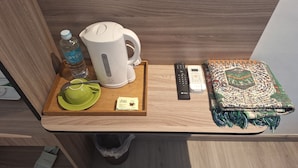 A wooden tray with a kettle, remote controls, a water bottle, a green cup, a spoon, a tissue, and a folded blanket.