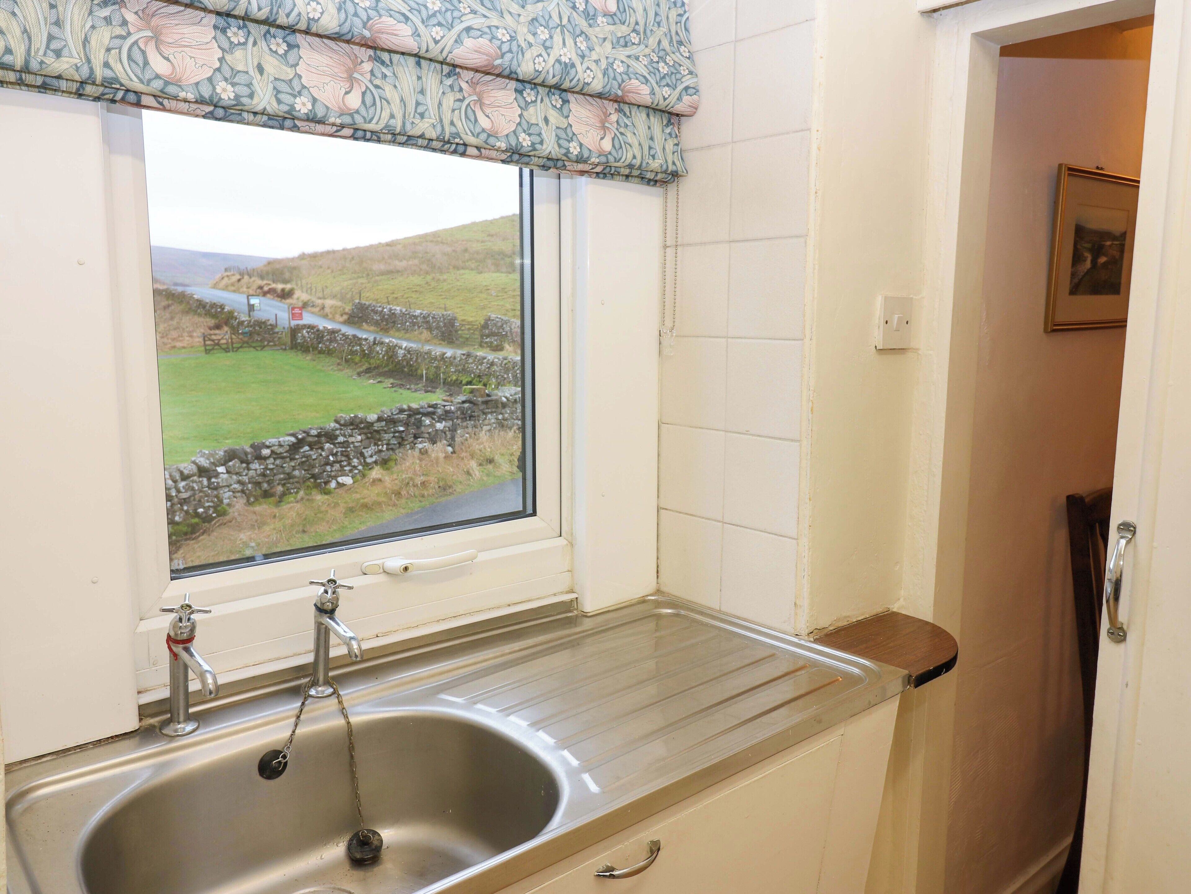 A kitchen with a sink, a window with a view of a stone wall and greenery, and a door leading to another room.