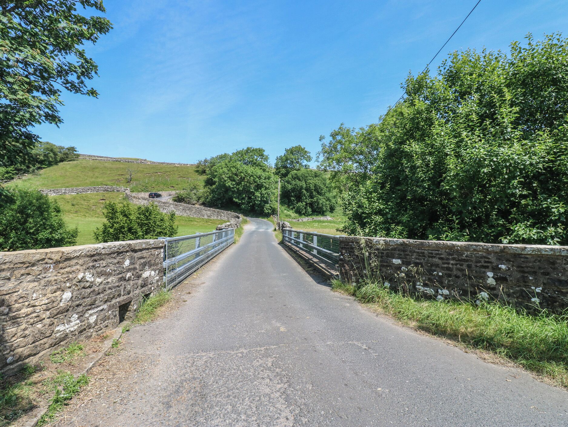A rural road with stone walls on either side, leading to a bridge over a small water body.