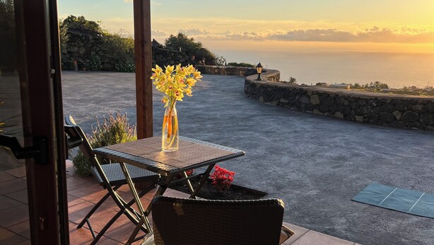 An outdoor patio with a table and chairs, a vase with flowers, and a view of the sea at sunset. - Peaceful Sea View Holiday Home with Stunning Sunsets". (Los Llanos De Aridane)