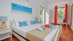 Un dormitorio con cama, mesita de noche, lámpara, un cuadro de playa, ventana con cortinas rojas y vista a zonas verdes.