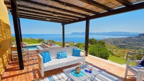 Un espacio de estar techado al aire libre con un sofá blanco y una mesa, con vistas panorámicas al mar y las colinas.