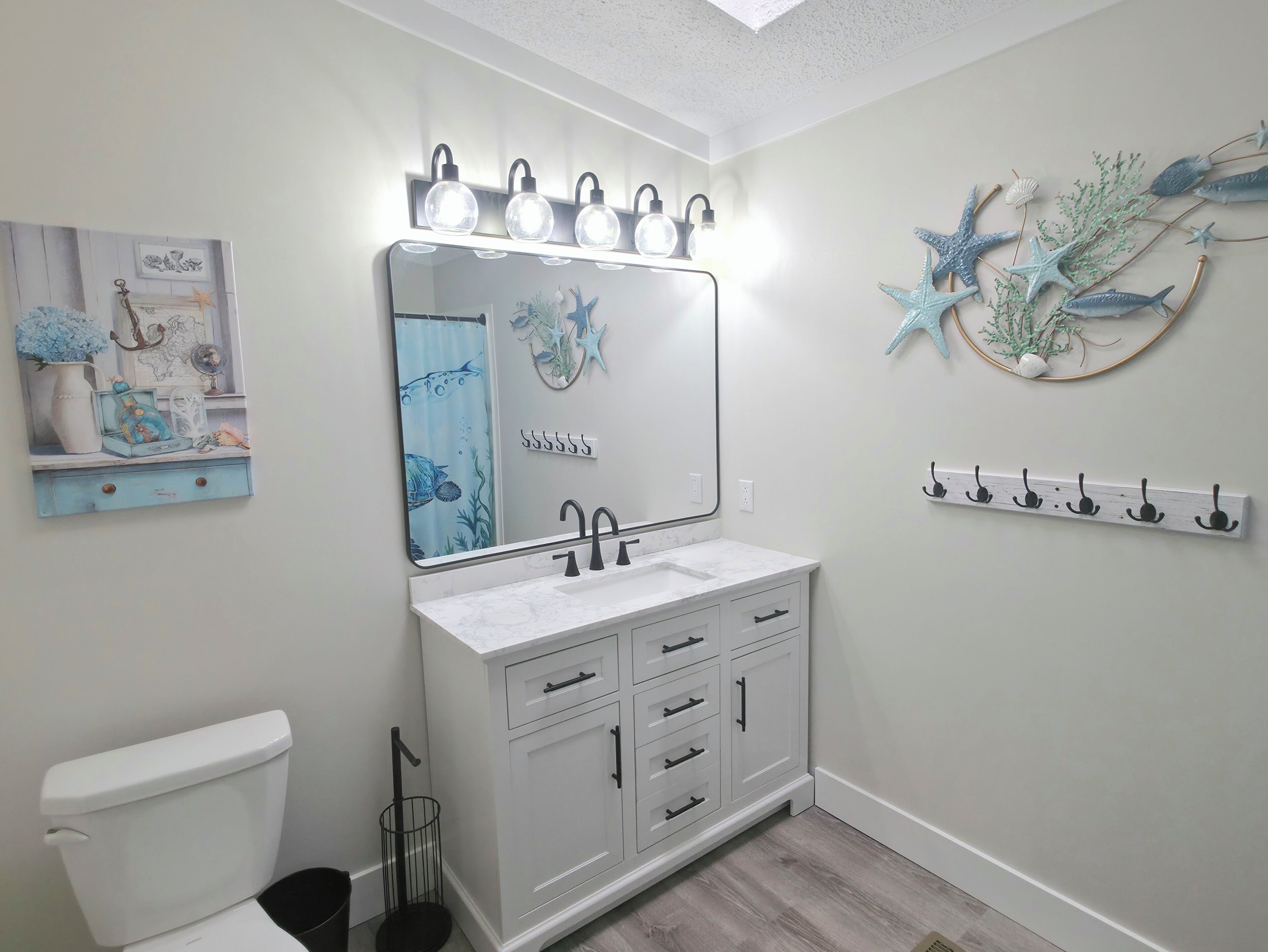 A bathroom with a white vanity, a large mirror with lights, a toilet, and a wall-mounted shelf with decorative items.