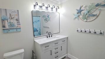 A bathroom with a white vanity, a large mirror with lights, a toilet, and a wall-mounted shelf with decorative items.