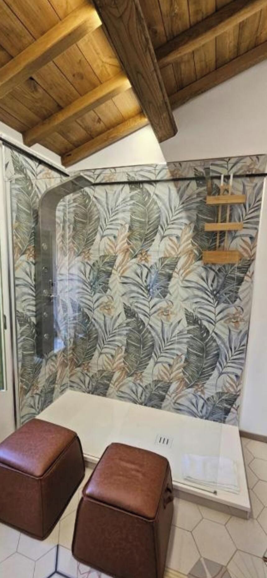 A shower with a glass door and a tropical leaf pattern. There are two brown stools in front of the shower.