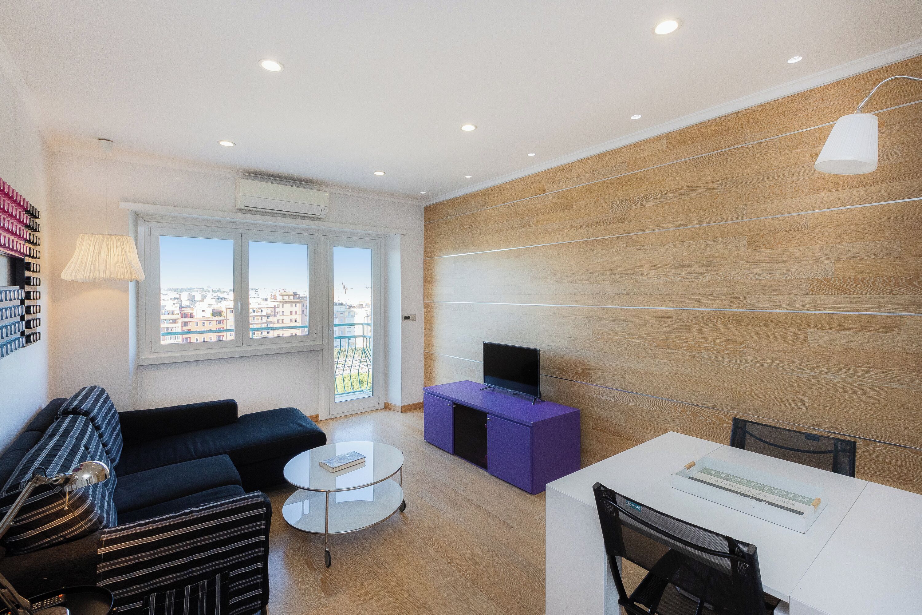 Un salon moderne avec un canapé, une table basse, un téléviseur posé sur un meuble violet et une vue sur la ville grâce à de grandes fenêtres.