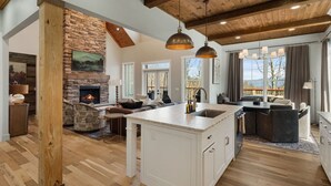 Un vaste espace de vie agrémenté d’une cheminée en pierre, de poutres apparentes et d’un grand îlot central doté d’un évier.