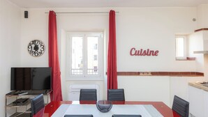 A dining area with a large table, four chairs, a TV, and a kitchen area with a red curtain. - Rental in Rome Principe Amedeo Apartment (Rome)