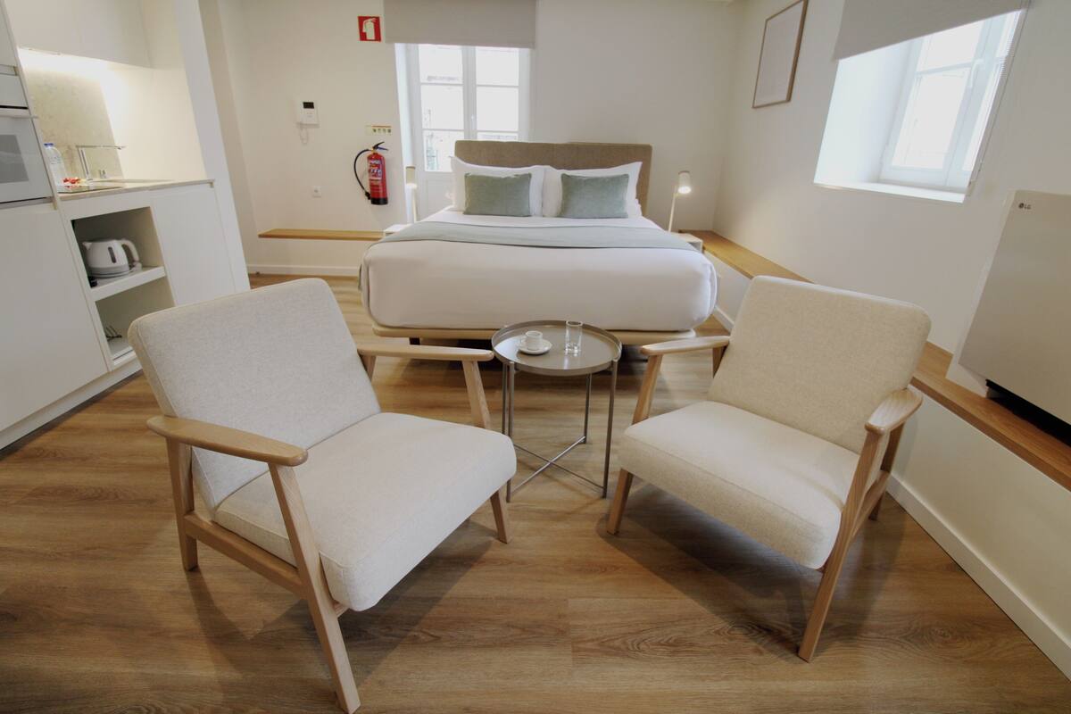 Une chambre d’hôtel moderne avec un grand lit, deux fauteuils, une petite table ronde et, au fond, un coin cuisine.